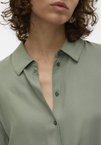 Chemise vert clair avec un col, dotée d'un devant à boutons, texture lisse et léger brillant. Les boutons sont de couleur contrastante foncée.
