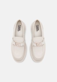 Mocassins en cuir beige avec un bout rond, des coutures tonales et un détail matériel en forme de "K" sur la sangle. Design lisse et minimaliste.