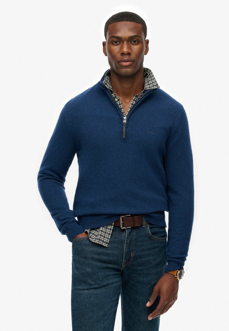 Sudadera de cremallera azul marino en una textura suave, superpuesta sobre una camisa de cuadros, combinada con jeans de mezclilla y un cinturón marrón.