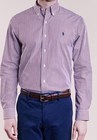 Långärmad knapp-up skjorta med smala vertikala lila och vita ränder, som har en liten blå logotyp på bröstet, matchad med mörkblå byxor.