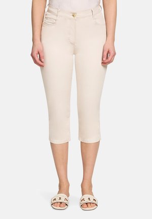 Kobieta w beżowych krótkich spodniach z przednim guzikiem i sandałach, stojąca na białym tle.