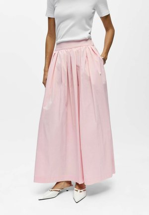 Donna che indossa una maglietta bianca a maniche corte, una lunga gonna plissettata rosa chiaro con tasche e scarpe bianche con tacco basso a punta e fiocchi.