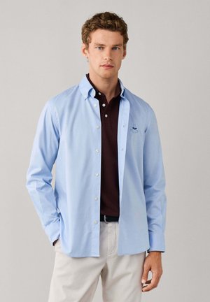 Joven con camisa azul claro abotonada sobre un polo marrón oscuro, de pie con las manos en los bolsillos de unos pantalones beige claro frente a un fondo liso.