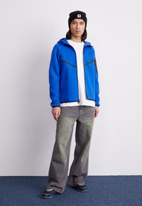 Sweatshirt azul com fecho e detalhes em preto, t-shirt branca, calças largas cinzentas com um acabamento desbotado, e um boné preto. Sapatos pretos texturizados.