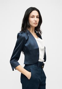 Femme aux longs cheveux foncés portant un blazer court bleu marine, un chemisier blanc et un pantalon taille haute bleu marine, les mains dans les poches, sur un fond uni.