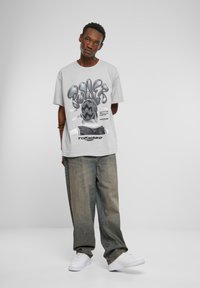 Gray graphic t-shirt featuring bold black text and a monochrome design on the back, paired with loose-fitting light denim jeans and white sneakers.