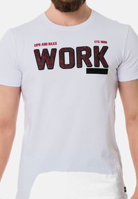 Weißes Baumwoll-T-Shirt mit fetten schwarzen Buchstaben, die "WORK" buchstabieren, und einem rot gesprenkelten Akzent. Rundhalsausschnitt und kurze Ärmel.