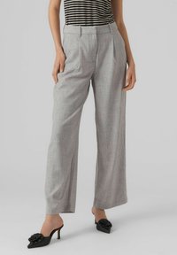 Pantalons larges gris avec un motif à carreaux subtil, comportant des plis à l'avant, taille haute, en tissu texturé, associés à des escarpins à talon ouvert noirs avec un accent floral.