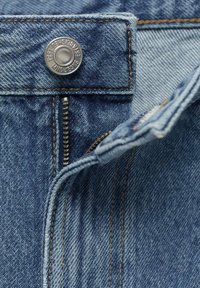 Blå denimtyg med synlig söm, en metallknapp i midjan och en metalldragkedja framtill.