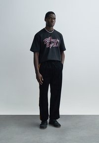 Μαύρο μπλουζάκι με το λογότυπο "Billionaire Boys Club" σε πολυχρωμία, σε συνδυασμό με μαύρο παντελόνι φόρμας με κίτρινες πλευρικές ρίγες και μαύρα παπούτσια.