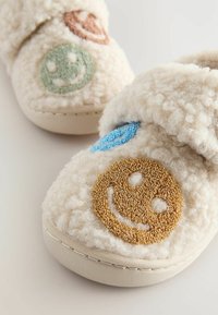 Chaussons en crème peluche avec des visages souriants colorés brodés en vert, bleu et marron. Texture douce et durable, semelle de couleur claire.