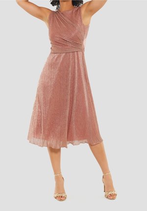 Robe sans manches en rose doré, longue jusqu'aux genoux, avec un design croisé, tissu texturé et jupe fluide. Associée à des sandales à talons métalliques.