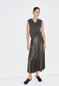 Šedý bezrukávový pletený svetr, olivově zelená skládaná midi sukně a černé dřeváky se stříbrnými akcenty. Minimalistický design a hladká textura.