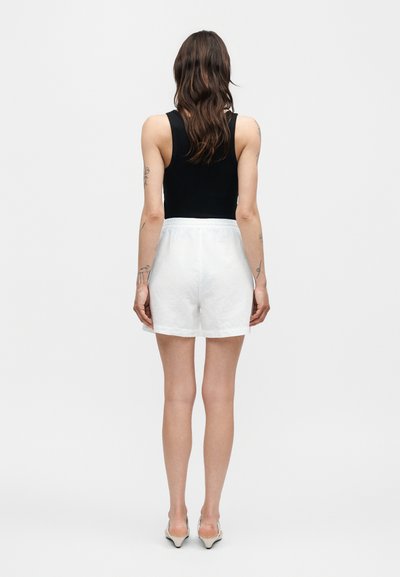 Γυναίκα με κυματιστά καστανά μαλλιά που φοράει μαύρο αμάνικο τοπ, άσπρα σορτς και μπεζ ψηλοτάκουνα, στέκεται με την πλάτη σε λευκό φόντο.