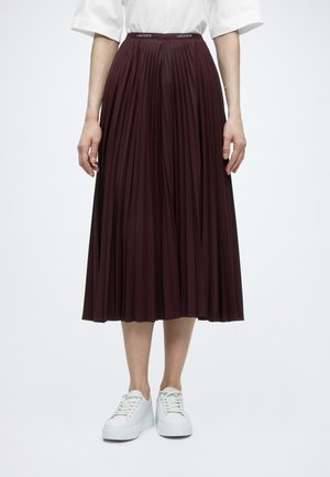 Femme portant un haut blanc, une jupe midi plissée violet foncé avec une ceinture au logo Lacoste et des baskets blanches, debout devant un fond uni.