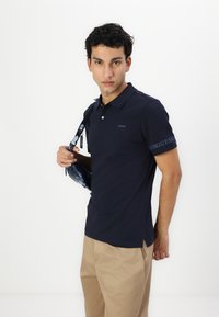 Cămașă polo bleu marin cu guler, mâneci scurte și un logo pe piept. Pur și simplu cu pantaloni bej și o geantă de umăr.