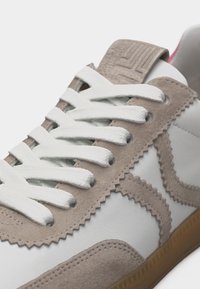 Κοντινή λήψη λευκού αθλητικού παπουτσιού με μπεζ σουέντ επενδύσεις, λευκά κορδόνια, λεπτομέρειες με ζιγκ ζαγκ στις άκρες και καφέ λάστιχο στη σόλα.