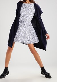 Mujer vestida con un vestido blanco de estampado paisley y cintura ajustada, cárdigan abierto azul marino, calcetines negros y mocasines negros con borlas.