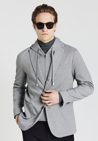 Giovane uomo che indossa occhiali da sole neri, felpa grigia con zip sotto un blazer grigio, dolcevita scuro e pantaloni neri, mentre con una mano aggiusta il blazer.