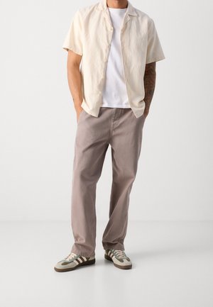 Man wearing a short-sleeve beige button-up shirt over a white T-shirt, light brown pants, and beige and green sneakers standing with hands in pockets.