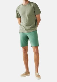 T-shirt verde oliva con tasca sul petto, abbinata a pantaloni corti verde chiaro e sneakers beige. Casual, materiale in cotone, maniche corte.