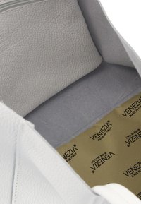 Das Innere einer weißen Lederhandtasche mit einer glatten, grauen Wildlederauskleidung und einem beigen Stoffboden, der ein sich wiederholendes Logo-Muster aufweist.