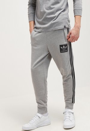 Person trägt graue Adidas-Sweatpants mit schwarzen Streifen und Logo, kombiniert mit weißen Sneakers und grauem Langarmshirt, steht mit den Händen in den Taschen.