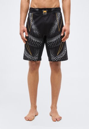 Άνδρας που φοράει μαύρο σορτς με γκρι σχέδιο φολίδων φιδιού και λεπτές κίτρινες γραμμές, όρθιος ξυπόλητος μπροστά σε απλό φόντο.