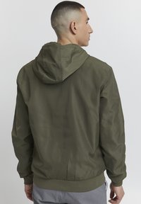 Veste à capuche vert olive avec une texture légère et lisse et des poignets élastiques. Dispose d'une taille ajustée et d'un design minimaliste.