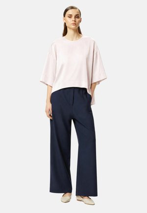 Giovane donna che indossa un top corto di colore rosa chiaro, pantaloni larghi blu navy e scarpe basse beige, in piedi con una mano in tasca.
