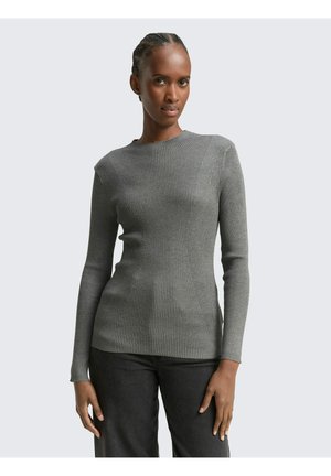 Frau mit geflochtenem Haar, die einen enganliegenden grauen Langarm-Rippstrickpullover und dunkle Hosen trägt, stehend vor einem schlichten hellen Hintergrund.