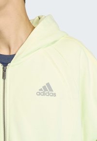 Svetlozeleni duks s kapuljačom i patentnim zatvaračem, s mekanom teksturom i sivim Adidas logotipom na prsima. Kapuljača djelomično preklopljena prema dolje.