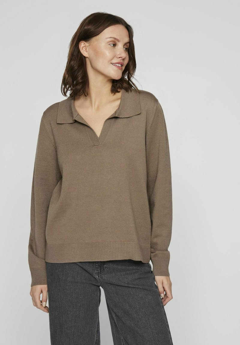 Suéter de punto marrón con cuello en V y solapas, puños y dobladillo acanalados, corte casual, combinado con pantalones de mezclilla de pierna ancha en gris oscuro, frente a un fondo neutro.