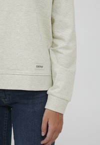 Hellgrau Sweatshirt mit strukturiertem Finish, einer frontalen Tasche und gerippten Bündchen. Mit einem OXMO-Label am Saum. Getragen mit dunkelblauen Jeans.