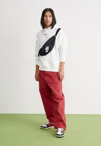 Borsa a tracolla nera in nylon con logo Nike bianco, indossata sopra un maglione con cappuccio bianco e abbinata a pantaloni rossi e sneakers nere.