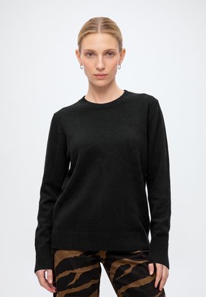 Vrouw met blond haar in een zwarte trui met lange mouwen en een broek met bruin-zwart patroon, staand tegen een effen lichte achtergrond.