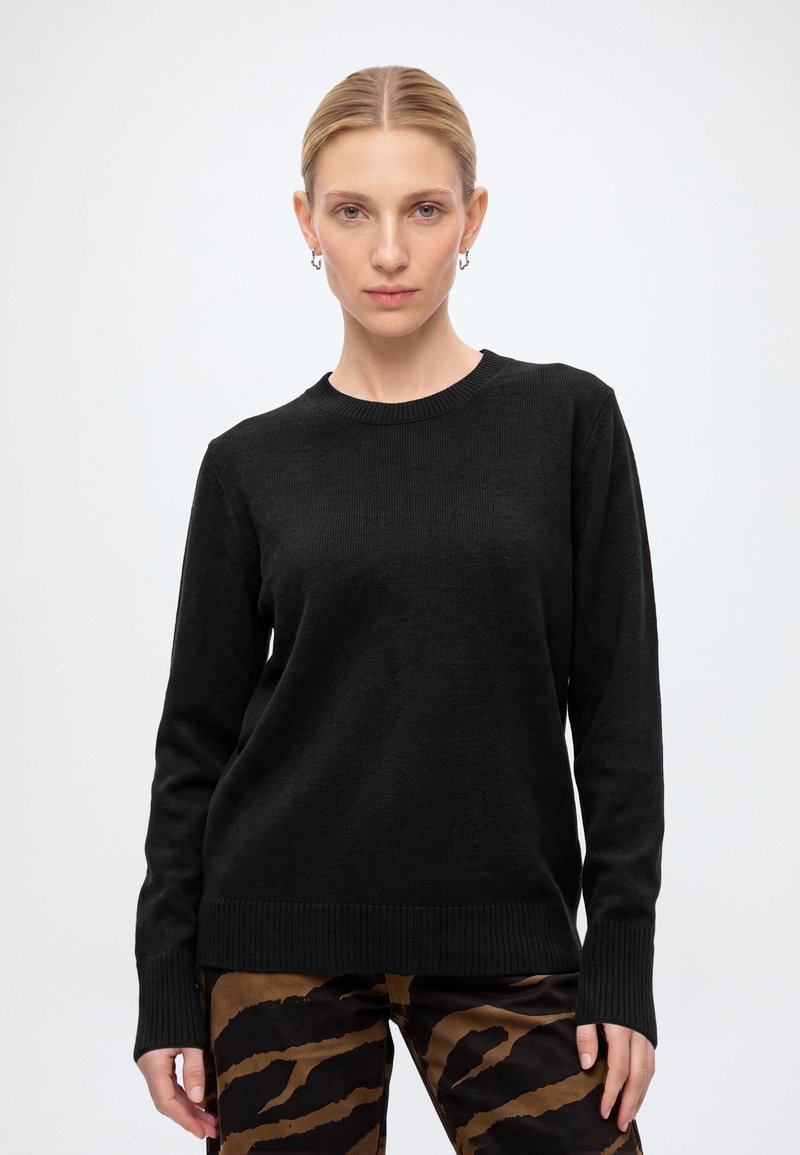 Femme aux cheveux blonds portant un pull noir à manches longues et un pantalon marron et noir à motifs, debout devant un fond clair uni.