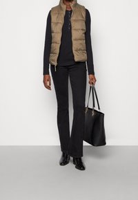 Gepolsterte olivgrüne Weste über einem schwarzen Langarmoberteil, kombiniert mit schwarzen Schlagjeans und schwarzen Ankle-Boots. Eine schwarze Handtasche mit Ketten rundet den Look ab.