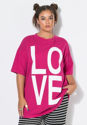 Woman with two buns hairstyle wearing a bright pink shirt with large white "LOVE" letters and black and white patterned pants.