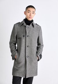 Grå hundtandsmönstrad kappa med hög krage, stora knappar och fickdetaljer. Mjuk textur, skräddarsydd passform och kontrasterande svarta detaljer.