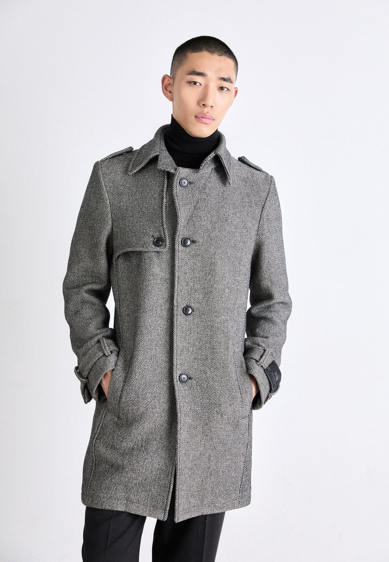 Grå hundtandsmönstrad kappa med hög krage, stora knappar och fickdetaljer. Mjuk textur, skräddarsydd passform och kontrasterande svarta detaljer.