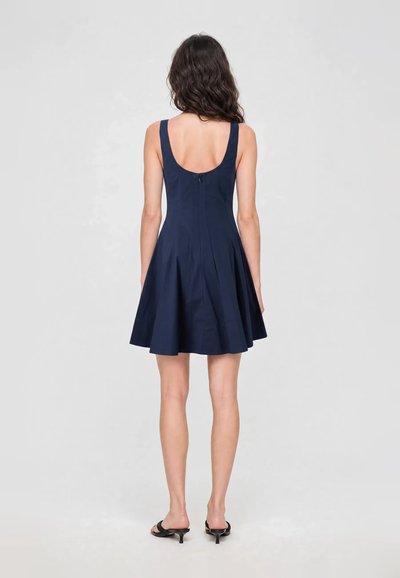 Femme aux cheveux ondulés et foncés portant une robe bleu marine sans manches en ligne A et des sandales noires à talons, debout, de dos, sur un fond uni.