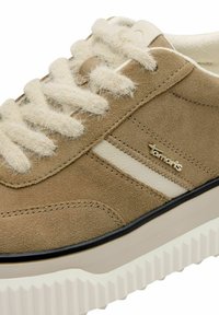 Tanfarbene Wildleder-Sneaker mit cremefarbenen Schnürsenkeln, erhöhter Gummisohle und goldfarbener Logoakzent. Merkmale: kontrastierende Seitenstreifen und weiche Textur.