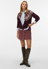Pull en tricot bordeaux avec motif de feuilles, blouse bleue claire en dessous, jupe en velours côtelé rose avec des boutons, et bottes brunes jusqu'aux genoux.