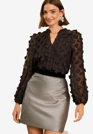 Blusa nera trasparente con accenti floreali testurizzati, caratterizzata da un collo a V e maniche lunghe. Abbinata a una mini gonna argentata metallica.