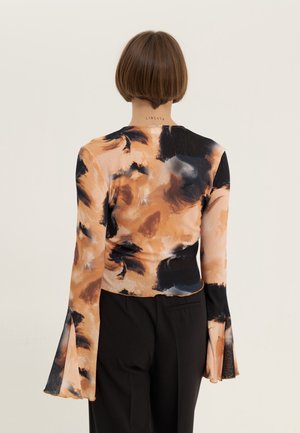 Vrouw met kort haar die een lange mouw draagt met een abstract zwart en beige patroon met uitlopende manchetten en zwarte broek, gezien van achteren.