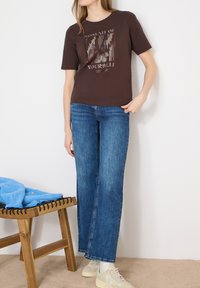 Femme portant un T-shirt graphique marron avec l'inscription "Nomad of Yourself", un jean bleu droit, des baskets blanches, debout à côté d'un banc en bois avec une veste bleue.