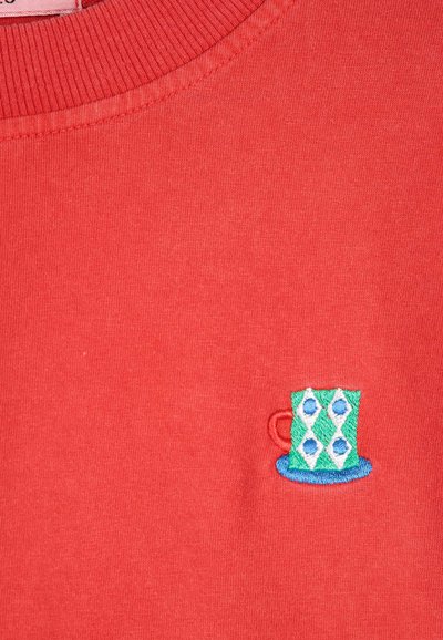Tissu rouge avec une petite tasse brodée verte et blanche avec des points bleus et une anse rouge sur une soucoupe bleue près de l'encolure.