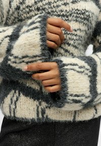 Sweter dzianinowy w kolorze kremowym i szarym, w luźnym fasonie, z puszystą teksturą, szerokimi rękawami i ściągaczem na końcu. Częściowo widoczne ręce.