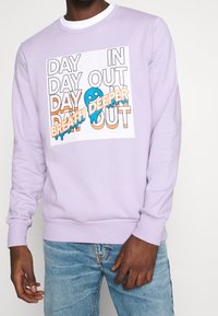 Lila sweatshirt med en vit grafik som har orange och blå text. Designen inkluderar orden "DAY IN DAY OUT" och en blå karaktär.